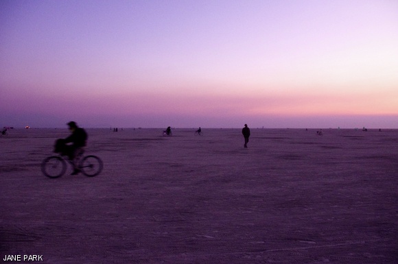 Burning Man participants walk and bike to and fro as the sun sets over the playa.