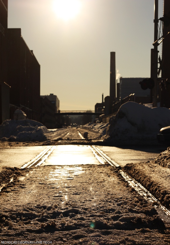 Railroad crossing on Mass. Ave. near Vassar Street, shot looking directly into the late afternoon sun. The Metropolitan Storage Warehouse is visible on the left; smokestacks from the Francis Bitter Magnet Lab stand on the right side of the picture. The strong backlighting provided by the sun silhouettes the buildings and is reflected in the exposed railroad tracks and the melting snow.
